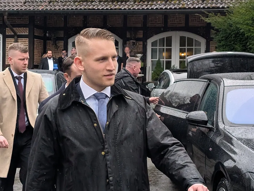 Das Foto zeigt Enrique Feldhaar am 22.02.26 vor dem Gasthaus "zur Post" in Billerbeck, wohin sich bei dem Besuch Björn Höckes die zugelassenen AfD-Gäste zurückzogen. Links hinter Felshaar in beigem Mantel ist Felix Hampe zu sehen. Feldhaat trägt die blonden Haare kurzrasiert mit einem strengen Seitenscheitel und eine dunkle Wachsjacke. Im Bildhintergrund rechts, vor der Fachwerkfassade des Gasthauses, ist Daniel Kokott zu sehen.