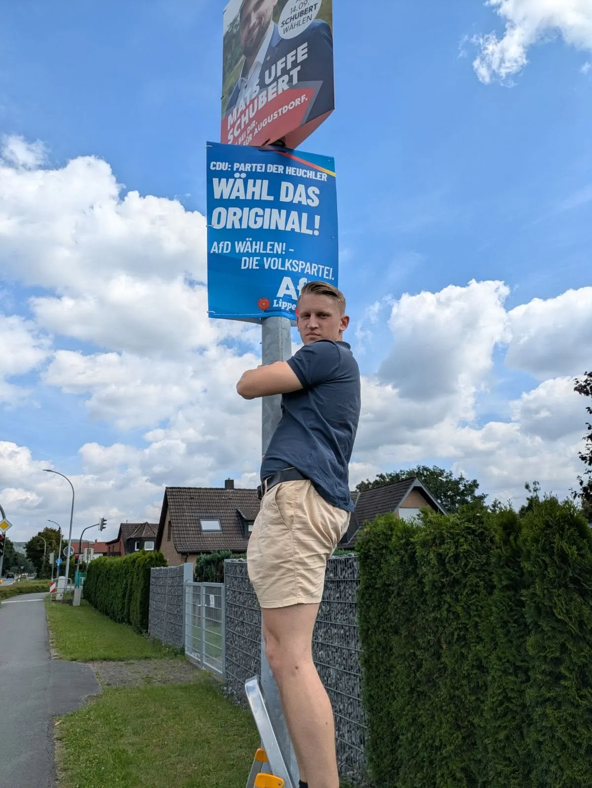 Das Foto zeigt Enrique Feldhaar auf einer Leiter stehend an einem Laternenpfahl, wo er ein Wahlplakat der AfD anbringt. Im Hintergrund ist eine kleine Straße zu sehen, an deren rechtem Rand eine Hecke und Häuser zu sehen sind.